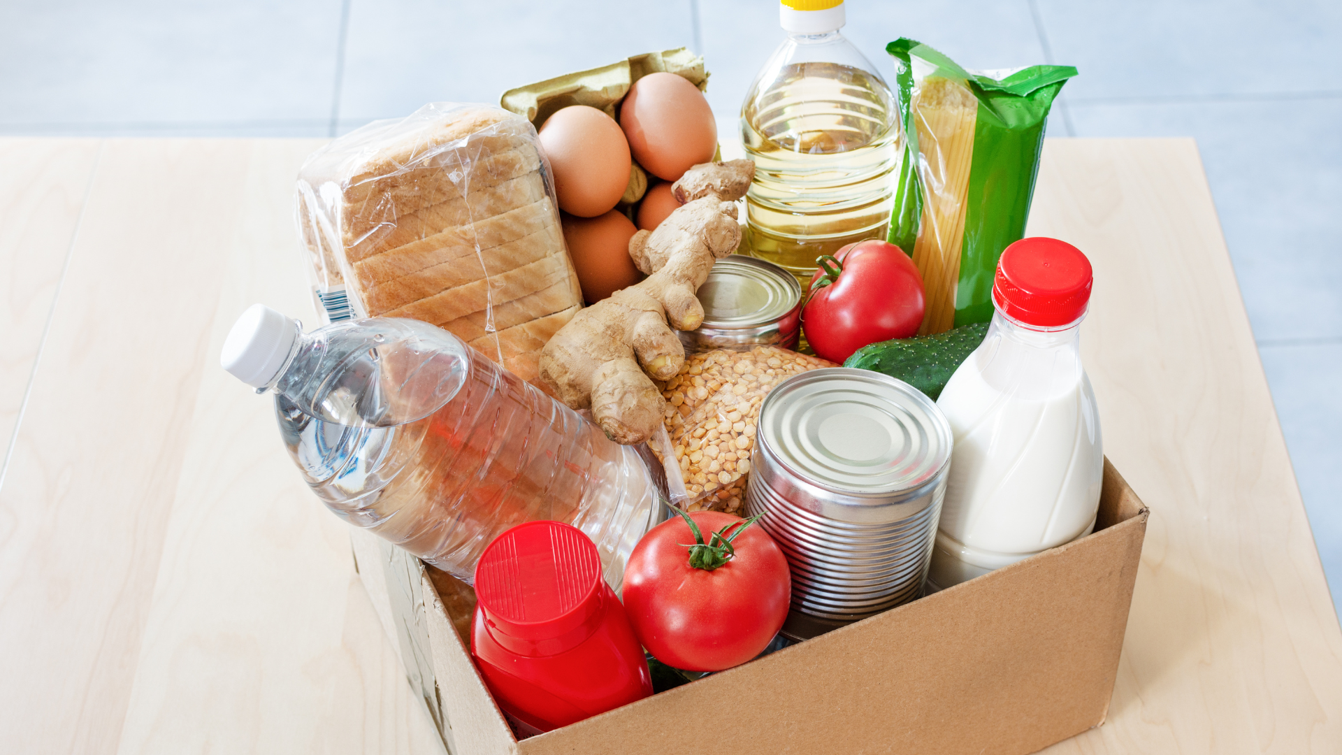 A cardboard box filled with food items, including milk, eggs, bread, ginger and tomatoes.