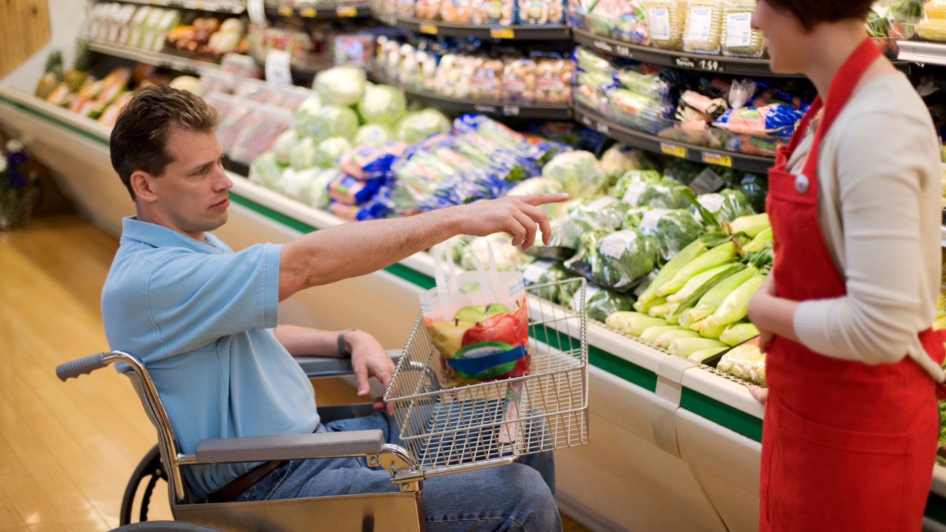 A person in a wheelchair points towards fresh produce in a grocery store, while a staff member in a red apron listens.