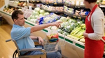 A person in a wheelchair points towards fresh produce in a grocery store, while a staff member in a red apron listens.