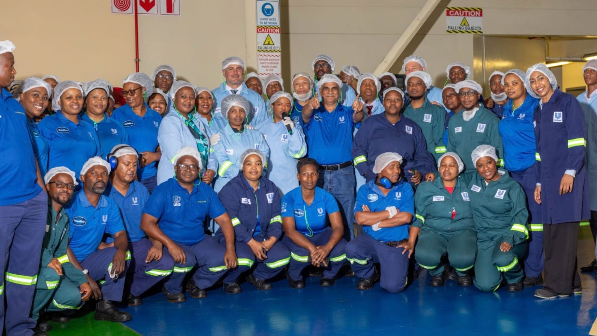 Factory employees in uniforms and hairnets pose together on the production floor.