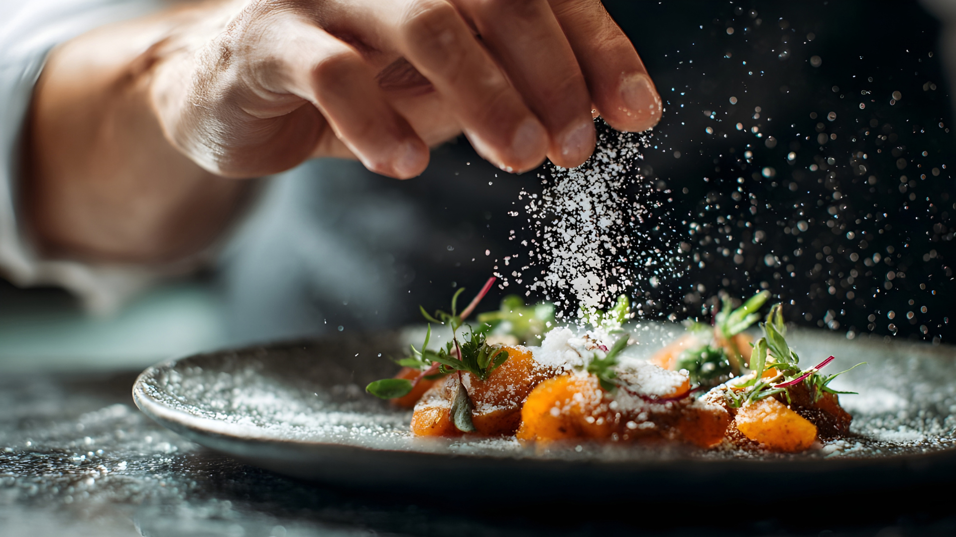 Chef sprinkling salt on gourmet dish with herbs on a dark plate.