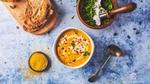 A bowl of orange soup, a small bowl of orange microalgae powder, a bowl of green herbs and a board with sliced brown bread.