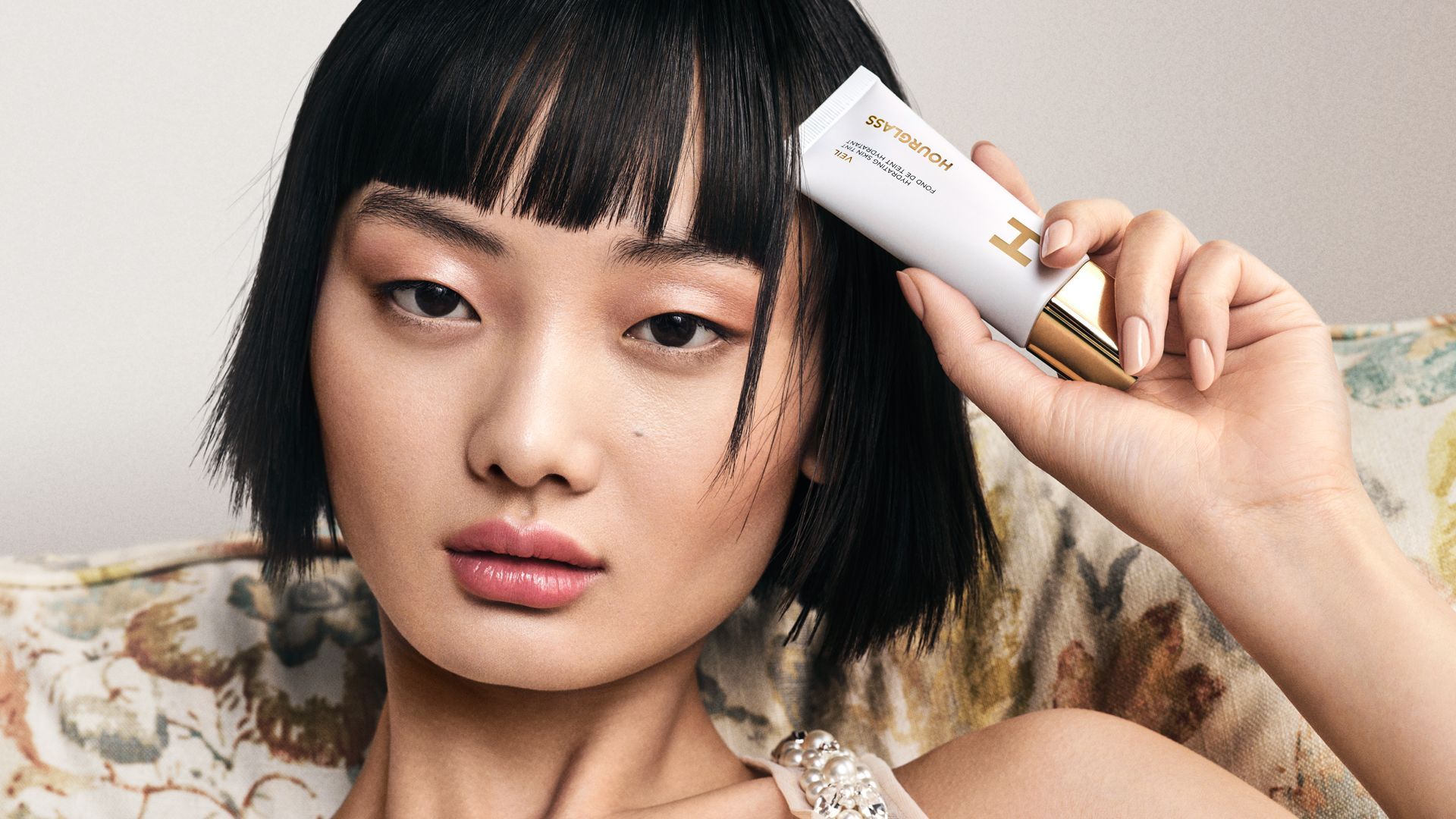 A women sat on a floral chair holding a white bottle of Veil by Hourglass by her head.