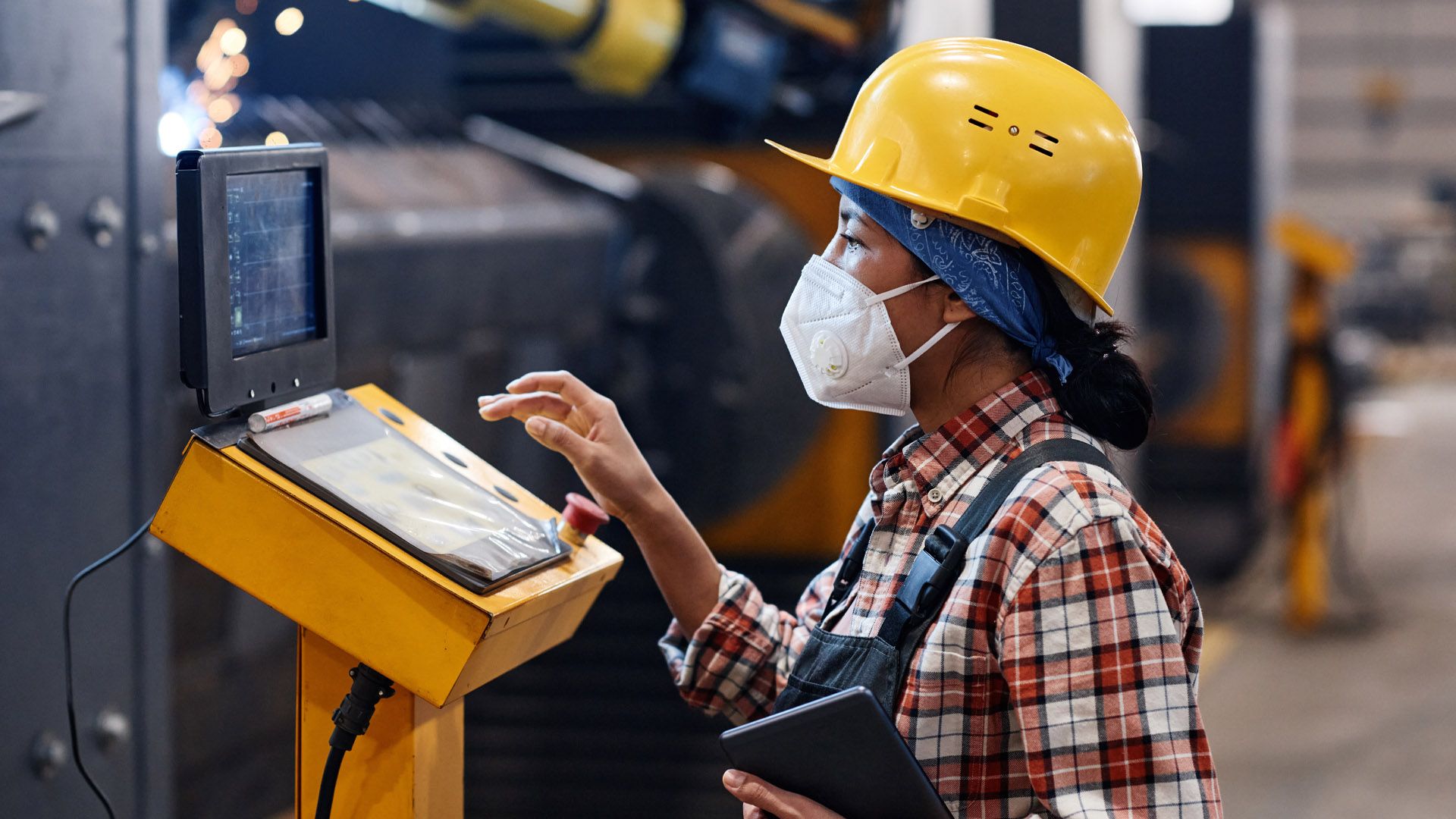 Woman factory worker in protective helmet, mask and coveralls operating a machine via a control panel.