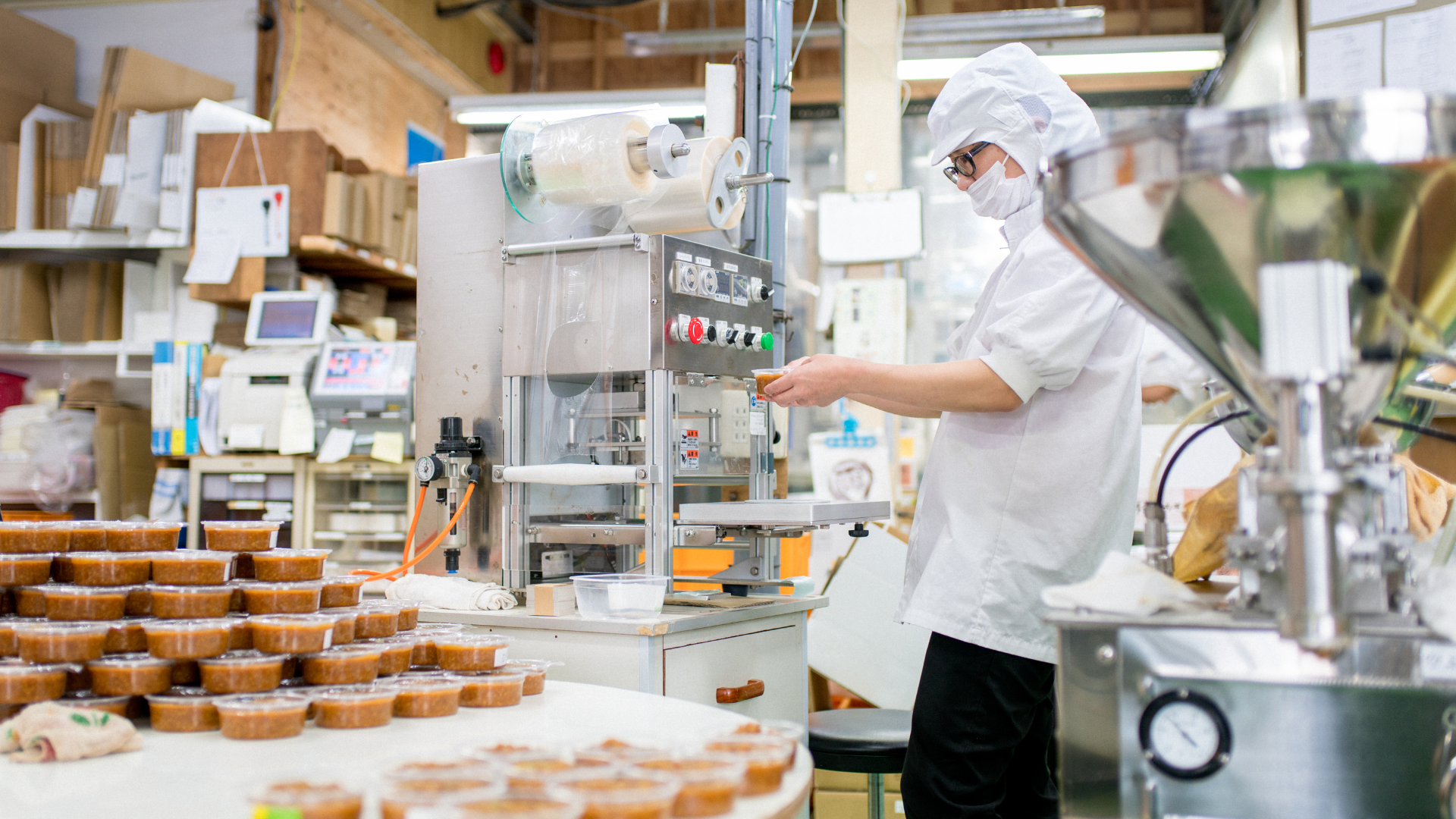 Worker in a factory using machinery with stacked food containers nearby.