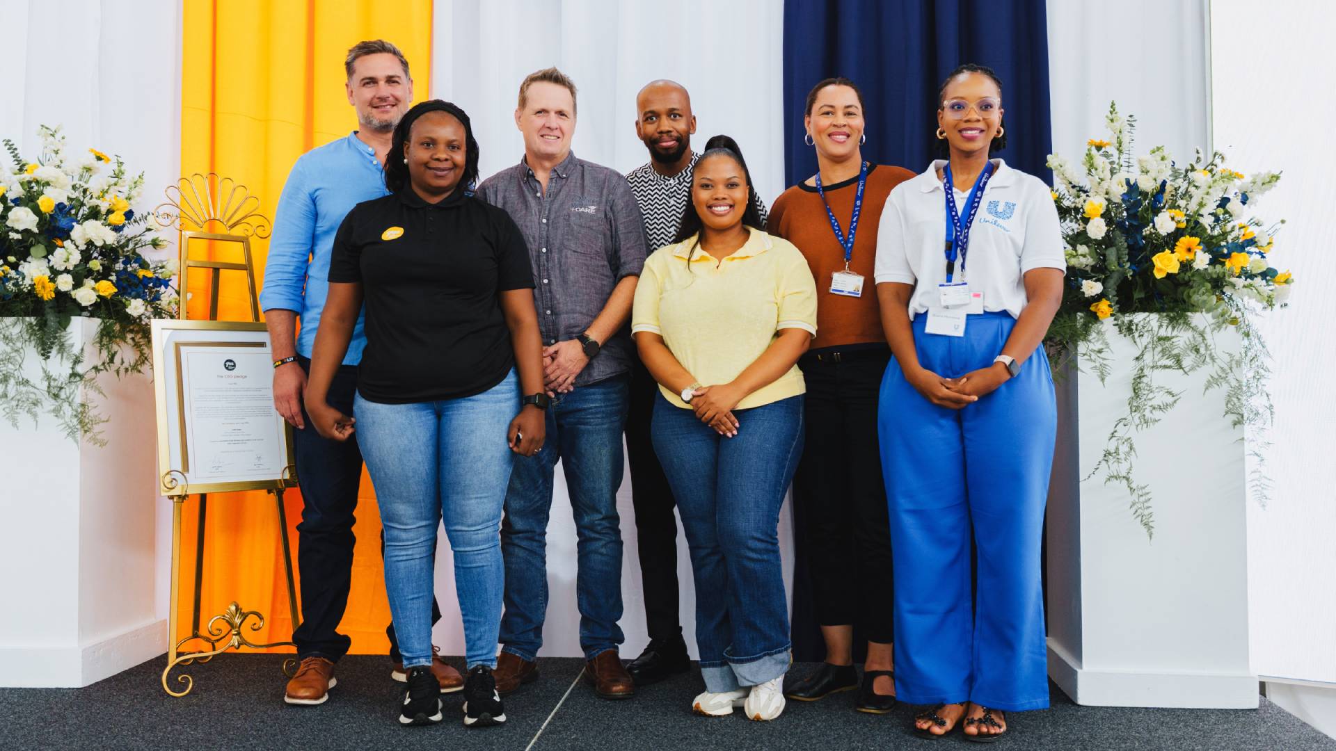 Group of seven people standing in front of floral arrangements and certificate