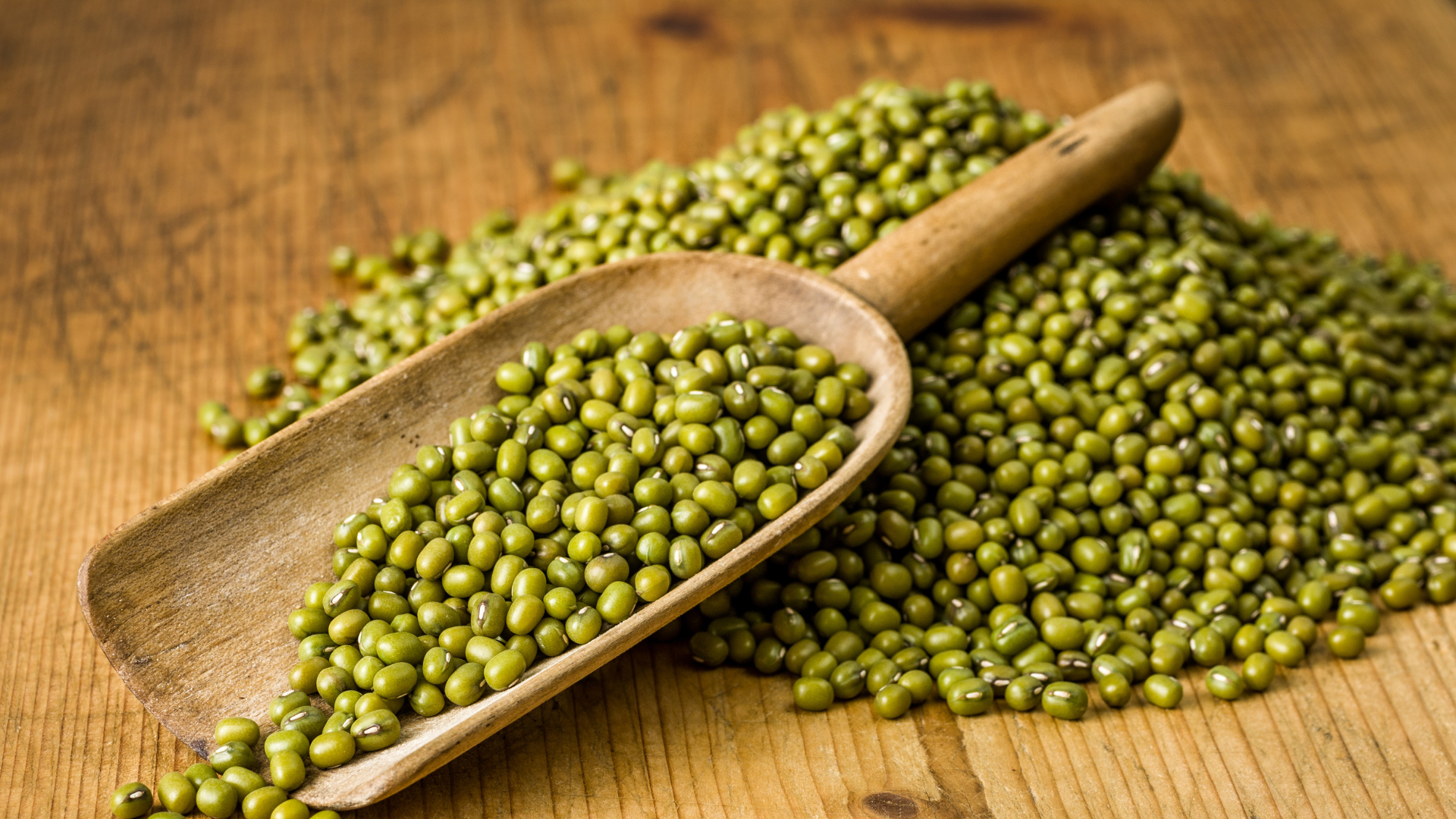 Wooden scoop with green mung beans on wooden surface.