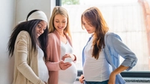 Three pregnant women stand together, smiling and looking at a smartphone.
