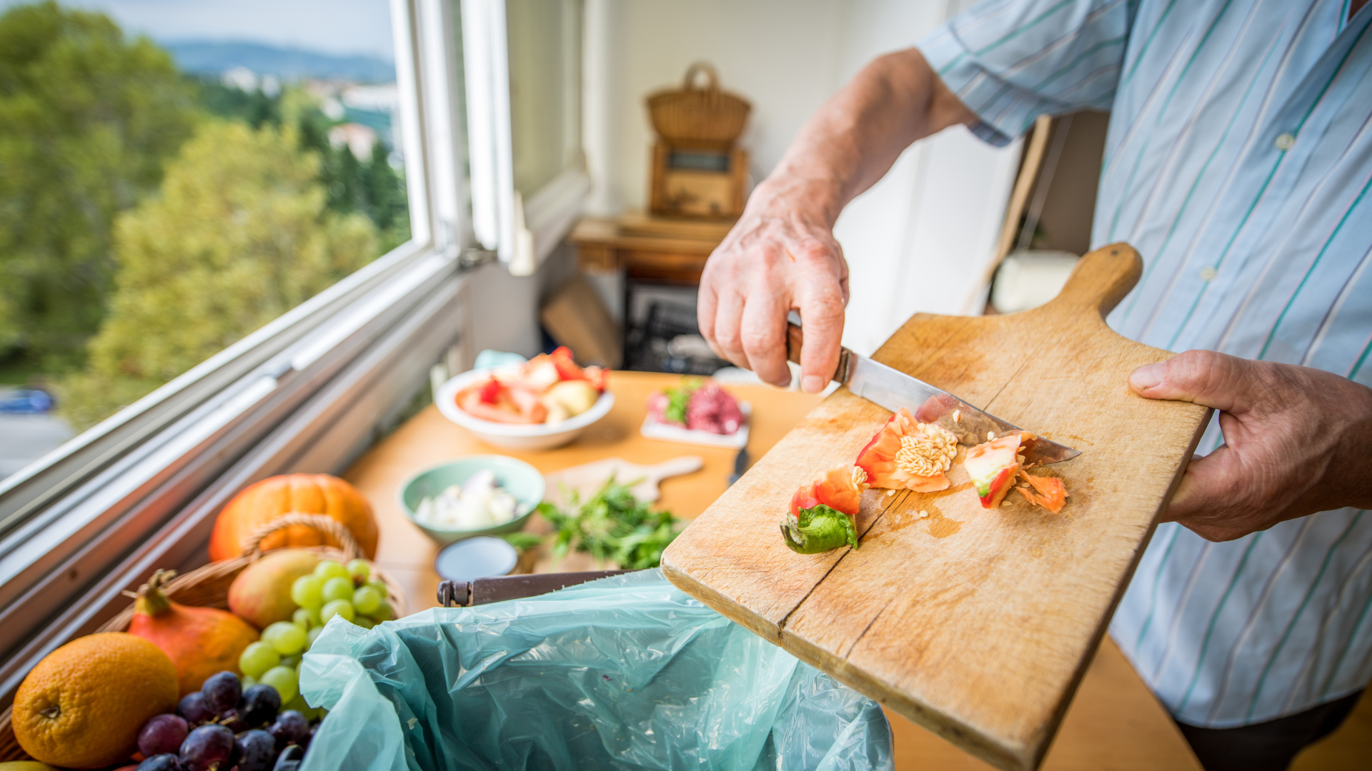 A person slices vegetables on a wooden cutting board, with a green trash bag nearby for food scraps and various fruits on the table.