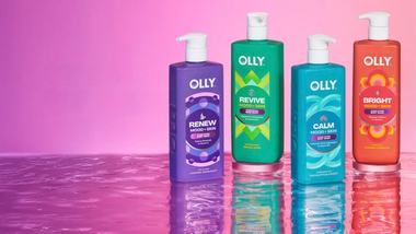 Four colourful Olly Mood + Skin body wash bottles stand on a reflective surface against a pink gradient background.