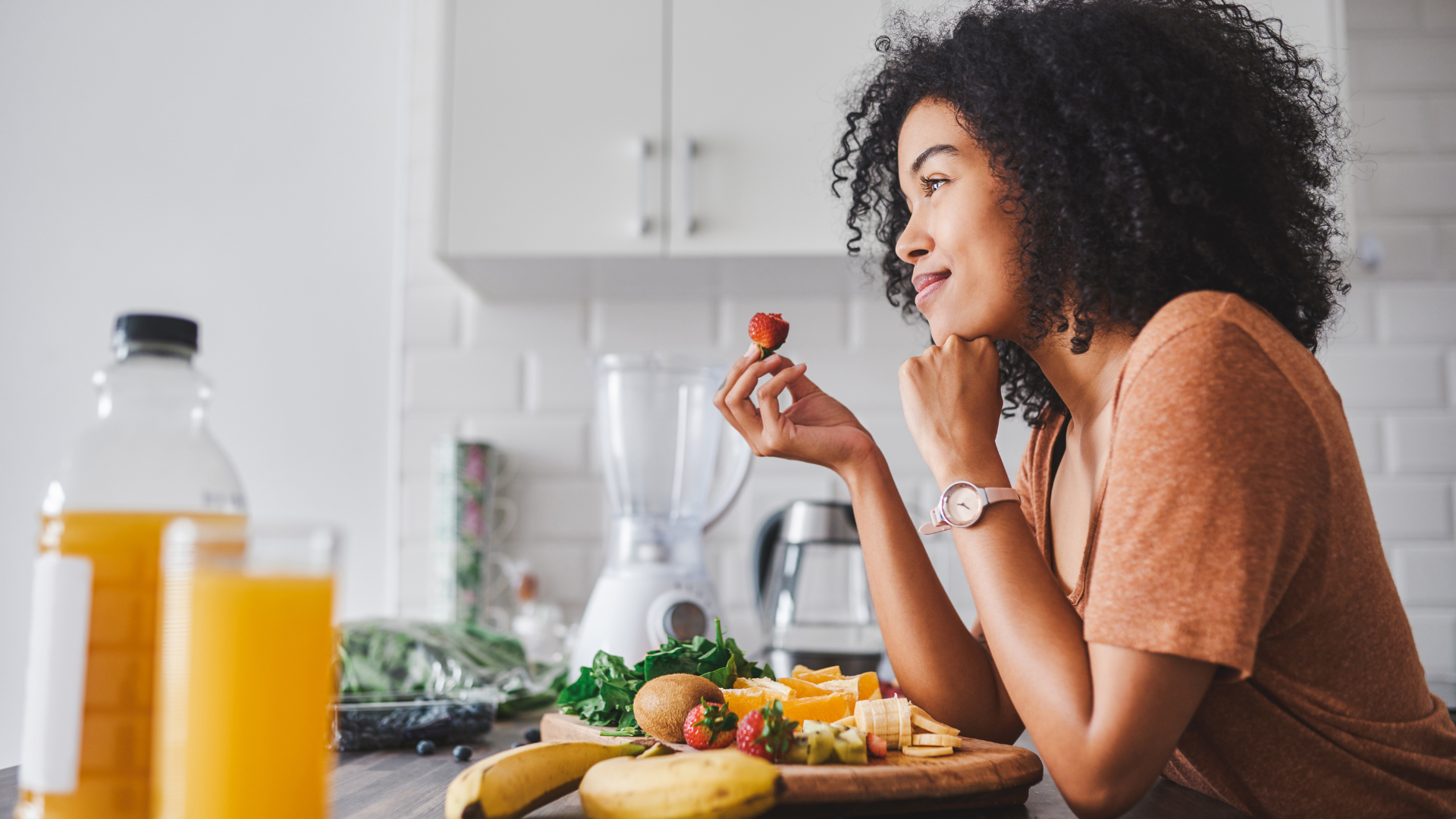 Woman enjoying fruit at kitchen counter with juice and blender.